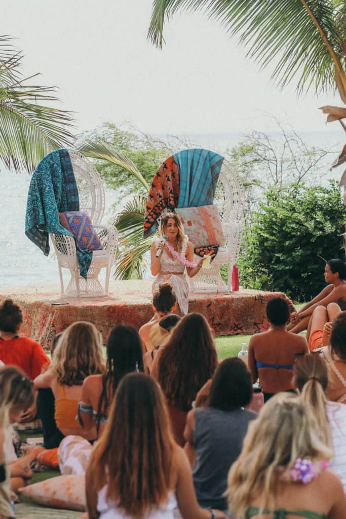 A retreat leader stands speaking to a group seated on the grass by the beach on Maui. Two wicker chairs with colorful drapes are behind her. Palm trees and the ocean provide a tropical backdrop, while the casually dressed attendees enjoy the outdoor setting.