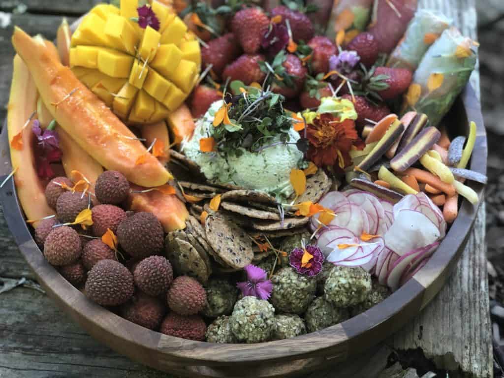 A colorful grazing board platter crafted by a Maui personal chef features sliced mango, papaya, lychees, strawberries, assorted crackers, cheese, edible flowers, radishes, carrots, and vegetable rolls. The artistic and vibrant arrangement is set on a wooden surface.