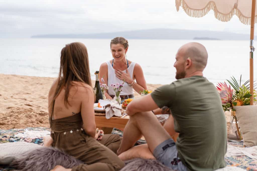 Three people sit on a beach under a parasol, enjoying a picnic on Maui from Lotus Chefs personal chef service. They are seated on blankets around a wooden table with snacks and drinks. The ocean and an island are visible in the background, creating a relaxed, sunny atmosphere.