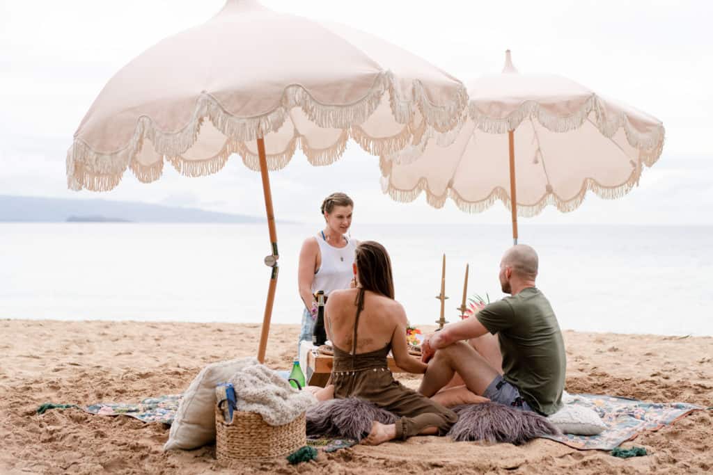 Three people sit on a beach under fringed umbrellas on a beach on Maui for a maui beach picnic. They are gathered around a small table with drinks and snacks on a colorful blanket. A basket with a blanket and pillows is nearby. The ocean is in the background.