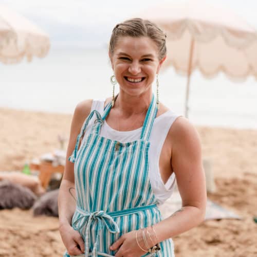 A person stands on a beach, smiling and wearing a blue and white striped apron over a white tank top. Chef Kyra Mirian Bramble from Lotus Chefs, known for her delectable creations, enjoys the coastal breeze with umbrellas and sand visible in the background.