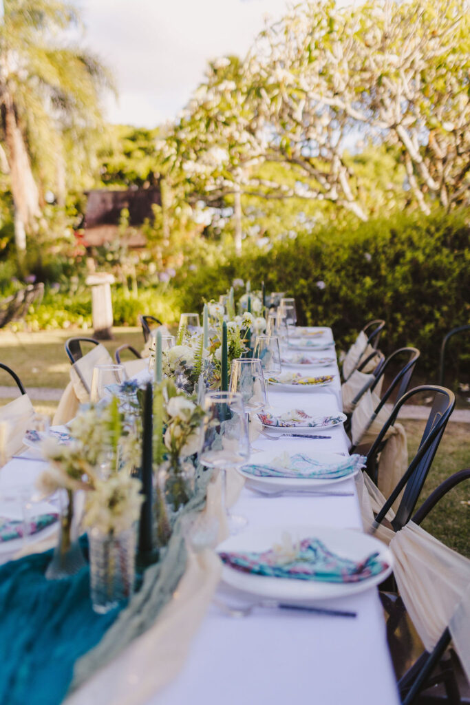 Outdoor dining table set for a private bridal party on Maui, surrounded by greenery. The table is elegantly decorated with a blue runner, tall candles, floral arrangements, and neatly arranged plates and glasses. Black chairs are placed along the sides.