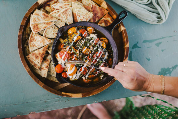 A hand holding a spoon over a skillet filled with roasted tomatoes, herbs, and cheese, surrounded by sliced pita bread on a wooden platter. The scene transports you to the vibrant cooking classes in Maui, all set against a teal surface. It is for a Maui yoga retreat catering job by a Maui private chef.