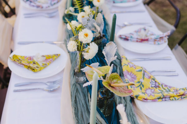 A beautifully set outdoor table with white plates, floral napkins, green candles, and a centerpiece of white flowers and greenery captures the essence of Maui private chef services, creating a fresh and elegant atmosphere perfect for an intimate gathering.