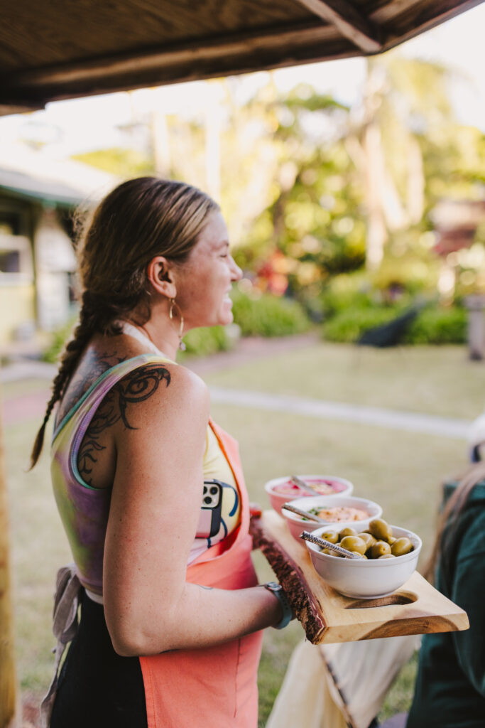 Chef Kyra Mirian Bramble from Lotus Chefs, with a braided hairstyle, wearing a colorful tank top and pink apron, is carrying a wooden serving tray loaded with small bowls of olives and other dishes. As they provide Maui private chef services, the backdrop of a house and lush greenery adds to the delightful scene.