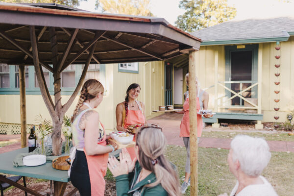 Four people wearing aprons chat and laugh under a wooden gazebo in a backyard, reminiscent of a Maui personal chef gathering. A house is in the background, and a table with food and drinks is nearby. In the foreground, two other individuals, one clapping, join this delightful scene.