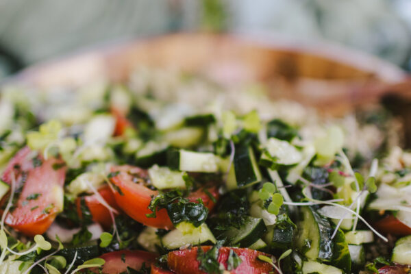 A close-up of a fresh salad crafted by a Maui personal chef, featuring sliced tomatoes, cucumbers, herbs, and microgreens atop a bed of quinoa in a wooden bowl, set against a blurred green background.