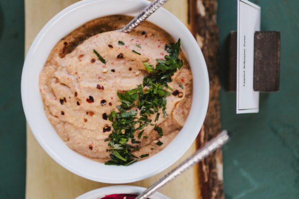 Three bowls on a wooden board: top with green olives, middle with a creamy dip topped with herbs and spices, bottom with a bright pink beet-based dip garnished with sesame seeds and oil. Perfect for your event when you hire Maui personal chef services.