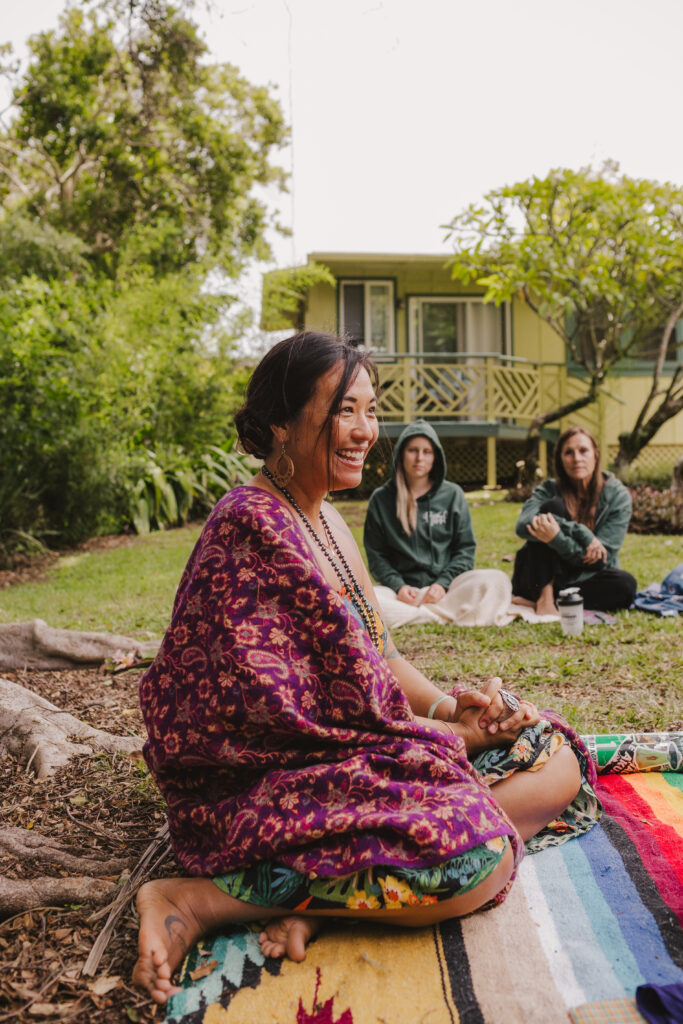 A woman sits cross-legged on a colorful blanket outdoors, wearing a patterned shawl and smiling. Nearby, two women relax on the grass, with trees and a house in the background. This serene setting echoes the tranquility found when enjoying Maui private chef services amid natures beauty.