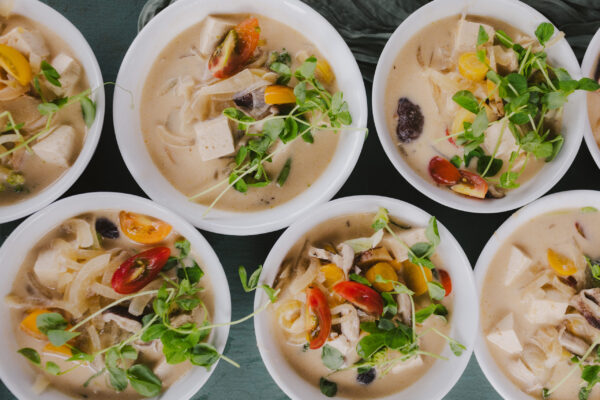 Bowls of creamy Thai soup garnished with microgreens, crafted by Maui personal chef for a plant-based retreat. The soup contains chunks of tofu, tomatoes, onions, mushrooms, and other vegetables, presented on a green surface.