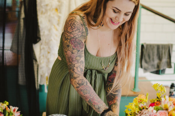 A woman with long hair and tattooed arms smiles as she arranges items on a table adorned with disco balls and colorful flowers, reminiscent of Mauis vibrant charm. Dressed in a green dress with a deep neckline, she embodies the artistry akin to Maui private chef services. Apparel hangs elegantly in the background.
