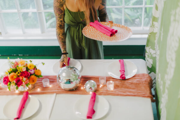 In a serene setting, a woman in a green dress—perhaps a Maui personal chef—sets a table with white plates and pink napkins. The table features a pink runner, colorful flowers, two disco balls, and small candles. Through the window, lush green foliage frames the scene.