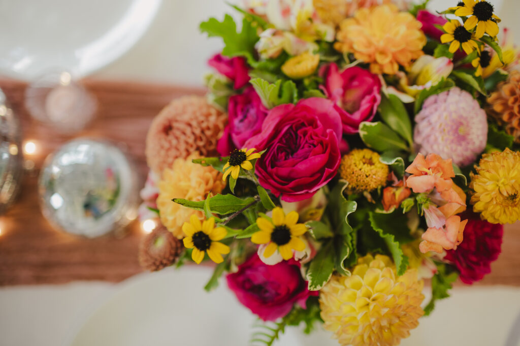 A vibrant bouquet including red roses, yellow mums, and small black-eyed Susans sits on a table with a blurred background, reminiscent of the elegance offered by Maui private chef services.