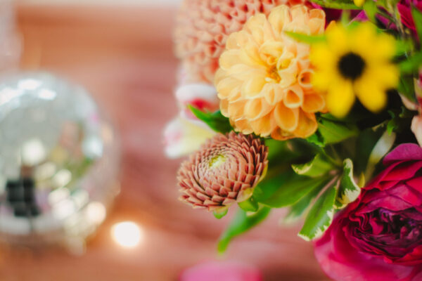 A vibrant floral arrangement with orange, yellow, and pink flowers graces the table, accentuating the elegance of a dining experience enhanced by Maui personal chef services. A pink napkin and white plate peek through in the foreground, while a silver orb-like decorative object sparkles in the background.