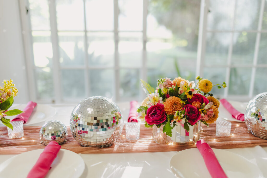 A table is decorated with colorful flowers in a vase, disco balls of various sizes, and pink napkins rolled on white plates. The background features bright, sunlit windows, creating a festive and elegant atmosphere perfect for enjoying a meal prepared by Maui private chef services for a lady brunch or bridal service.