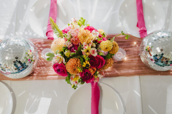 A vibrant floral centerpiece with pink, orange, and yellow blooms is surrounded by two disco balls on a table. Enhanced by Maui personal chef flair, the setting features white plates and pink napkins on a shimmering pink runner.
