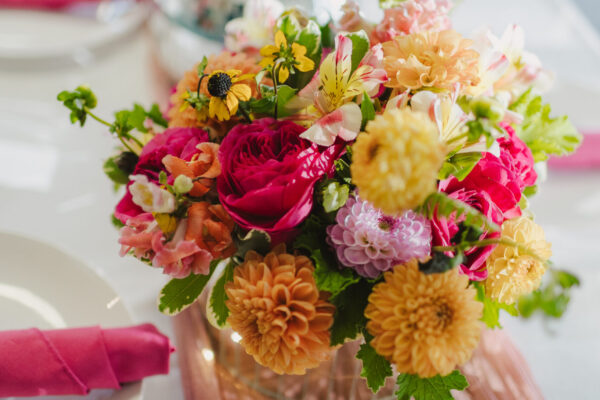 A vibrant floral arrangement featuring pink, orange, and yellow flowers in a glass vase graces the table, curated by a Maui personal chef for seated chef dinner or bridal service. Pink napkins are neatly rolled on white plates, complemented by a pink table runner and candle holder in the elegant setting.