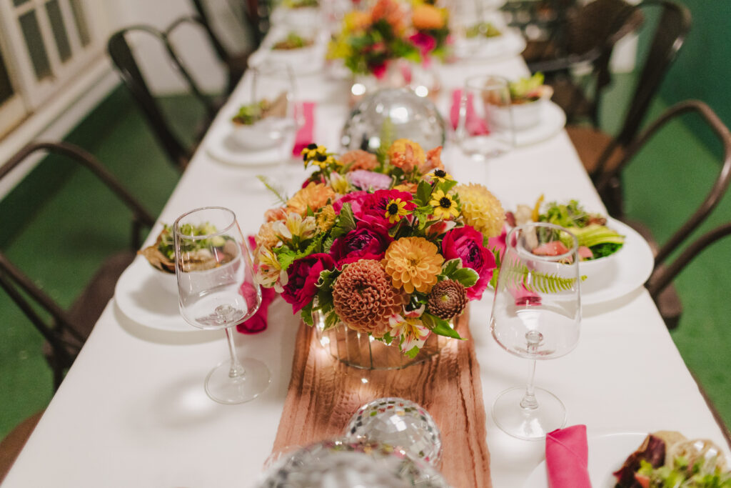 A long dining table is set with white linens, colorful floral centerpieces, and glassware. Plates hold salads beside pink napkins. With small silver disco balls adding a festive touch, the scene is perfect for an elegant gathering catered by Maui private chef services.