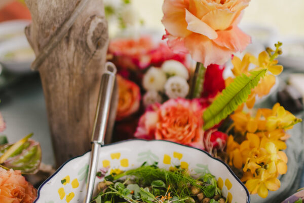 A colorful salad with greens and chickpeas is served in a decorative bowl, showcasing the fresh touch of Maui personal chef expertise for a retreat catering booking on Maui. The table is adorned with vibrant flowers in shades of orange, yellow, and pink. A utensil rests inside the bowl, and a wooden post is in the background.