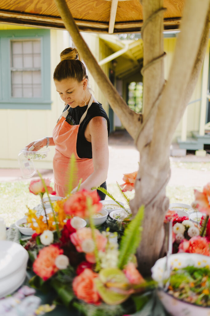 Chef Kyra Mirian Bramble from Lotus Chefs Maui wearing a pink apron stands behind a table, preparing food outdoors as part of Maui personal chef services. The table is adorned with colorful flowers and dishes. A tree provides partial shade, and a yellow building with green trim is visible in the background.