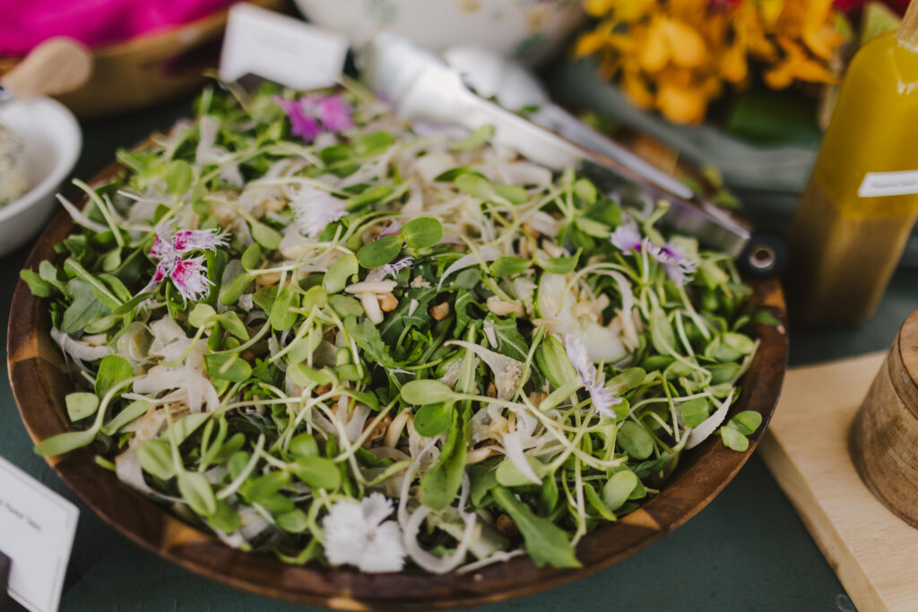 A wooden bowl brims with a fresh mixed salad of greens, sprouts, and edible flowers. A pair of tongs rests on the rim. In the background, colorful flowers and a bottle of dressing hint at the artistry typical of Maui private chef services for retreats and families by Lotus Chefs.