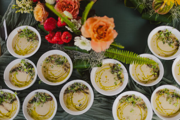 Bowls of cauliflower soup, expertly garnished with herbs and olive oil by a Maui based personal chef from Lotus Chefs, sit on a green tablecloth. Vibrant flower arrangements, including orange roses and red blossoms, are placed among the bowls, creating a decorative and appetizing display served by Maui private chef services.