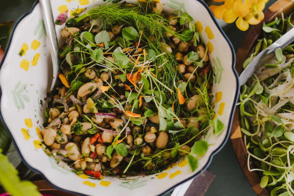 A bowl filled with a fresh salad of white beans, chopped herbs, and microgreens crafted by Maui private chef services. Garnished with edible flowers and surrounded by leafy greens, it boasts a decorative pattern with a spoon resting inside.