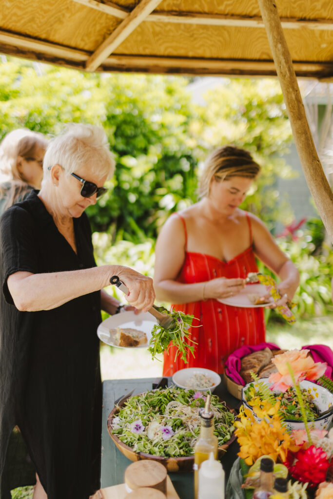 Two women serve themselves food from a buffet table filled with vibrant salads and colorful dishes, expertly crafted by Maui private chef services. They are outdoors under a canopy, surrounded by lush greenery. One woman is wearing sunglasses and a black dress; the other is in a red dress.