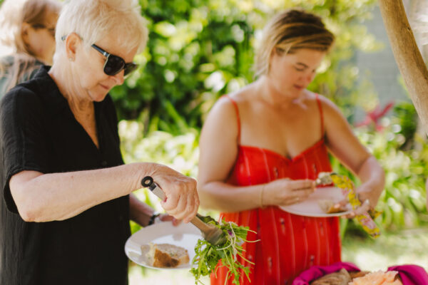 Two women serve themselves food from a buffet table filled with vibrant salads and colorful dishes, expertly crafted by Maui private chef services. They are outdoors under a canopy, surrounded by lush greenery. One woman is wearing sunglasses and a black dress; the other is in a red dress.