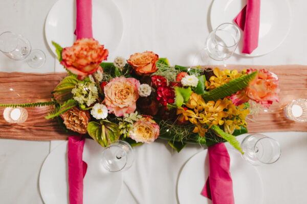A table set with a floral centerpiece featuring pink, orange, and yellow flowers evokes the charm of Maui personal chef dining. Pink napkins on white plates and clear water glasses enhance the ambiance. A light pink table runner and small candles complete this elegant arrangement.