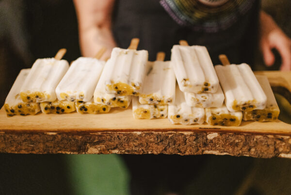 A wooden tray, reminiscent of a Maui private chefs touch, holds twelve creamy popsicles with visible chunks of passion fruit. The tray has a rustic bark edge, and the person holding it wears a dark shirt with a colorful circular design.