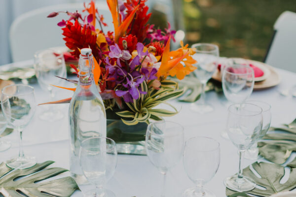 A round table set for an outdoor event on Maui, featuring a vibrant floral centerpiece with red, orange, and purple flowers. Plates with pink napkins, clear glasses, and tropical leaf decor are arranged neatly on a white tablecloth.