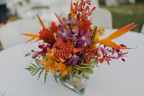 A vibrant floral arrangement in a clear vase featuring orange, red, purple, and yellow flowers with green fern leaves, placed on a white table. A floral arrangement for a private chef dinner service with tablescaping.