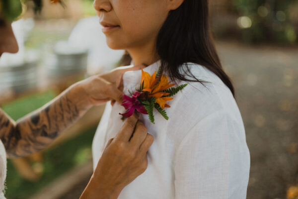 A bride in a white shirt is having a colorful flower boutonniere pinned by another with tattooed arms, creating a bridal styled moment. The vibrant orange, pink, and green flowers pop against the blurry outdoor backdrop. It is a perfect day for a lesbian wedding on Maui catered by Lotus Chefs!