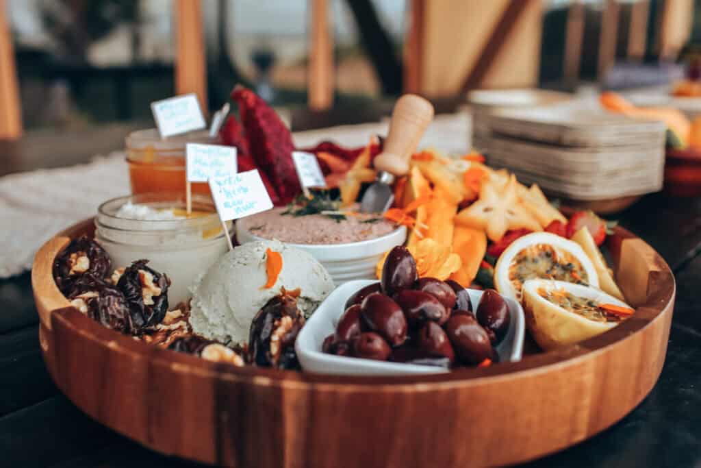 A wooden platter holds a variety of foods, including olives, cheese, fruit, and spreads for Maui pupu service. Small jars with labels and starfruit slices are also visible. The platter is set on a table with a blurred background.