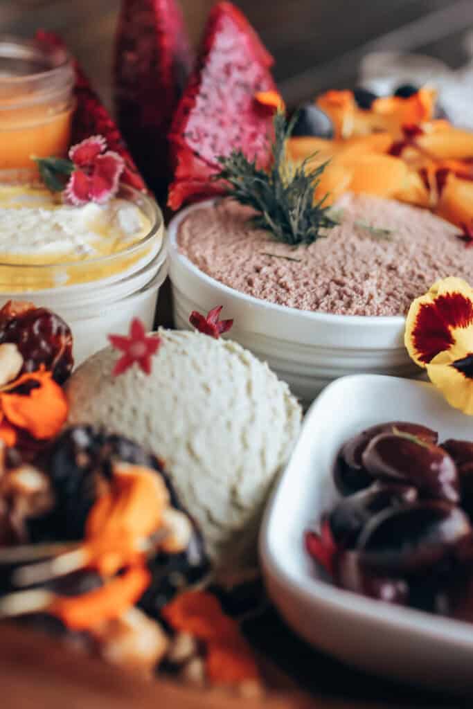 A close-up of a platter reminiscent of a Maui High Tea, featuring a variety of dips in bowls, garnished with fresh herbs and colorful edible flowers. Surrounding the dips are sliced fruits, nuts, and vibrant orange and red fruits, creating an elegant presentation.