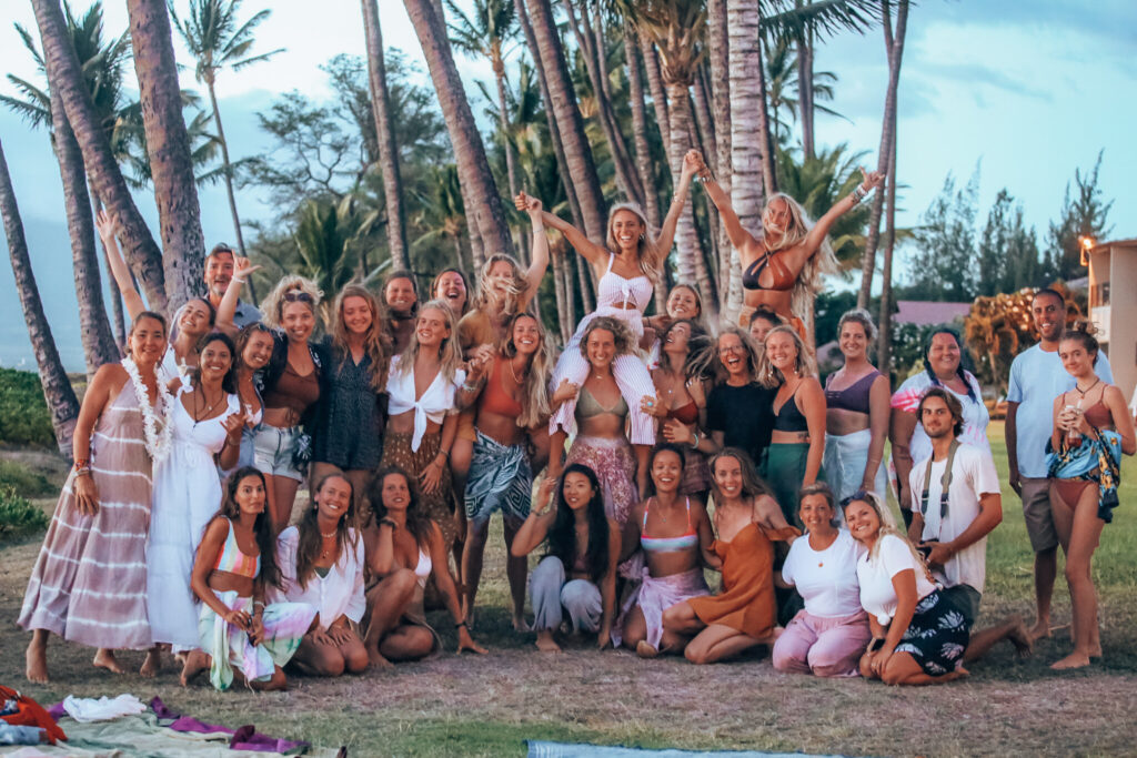 A large group of people, mostly women, pose happily outdoors with palm trees in the background at a retreat on Maui. They are dressed in casual, summery clothing, and some are raising their arms in celebration. The scene is relaxed and joyful.