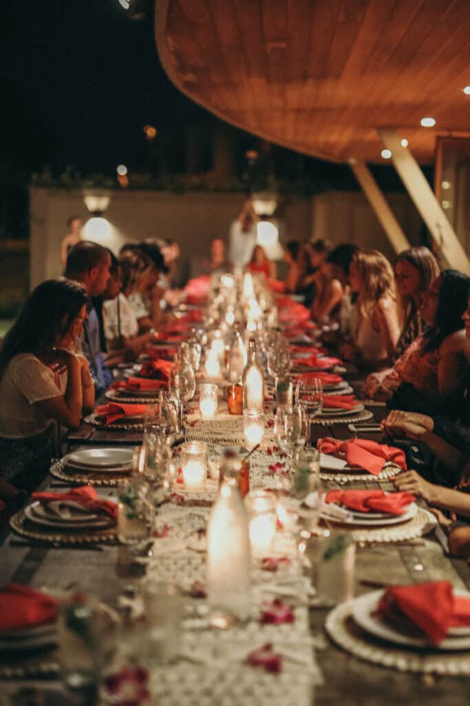 A long dinner table is set outdoors beneath a wooden ceiling. The table is adorned with red napkins, candles, and a white lace runner. Many people are seated around the table, engaged in conversation, creating a warm, elegant atmosphere while they wait for their private chef from Lotus Chefs.