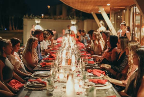 A large group of women sit around a long, elegantly set dining table adorned with candles and red napkins for a private dinner experience on Maui. The setting appears festive and intimate, with warm lighting creating a cozy atmosphere.
