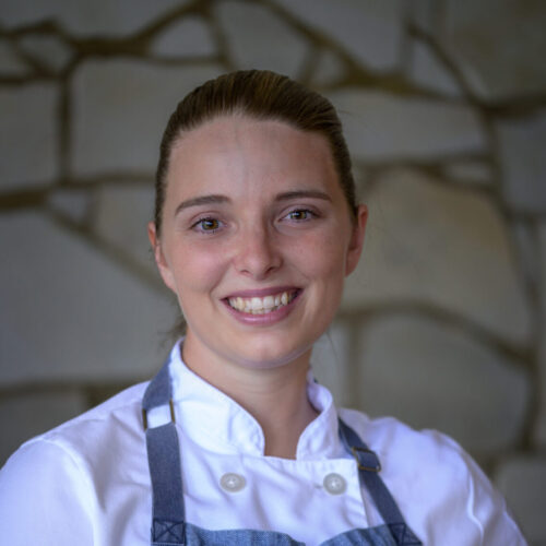 A smiling member of the Lotus Chefs team, Chef Alisha Rose Marquis, wears a chefs jacket and apron, standing confidently in front of a stone wall backdrop.