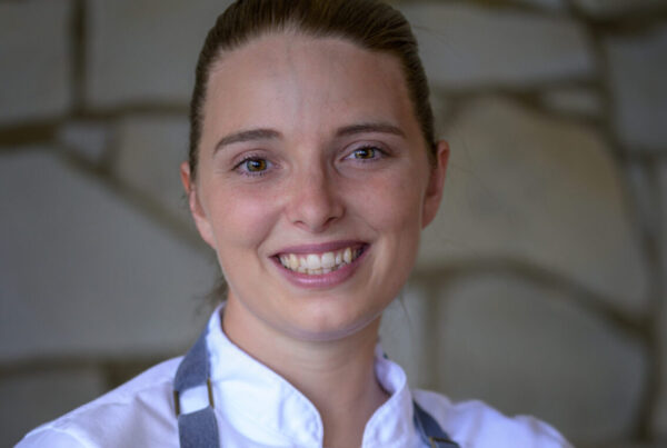 A smiling member of the Lotus Chefs team, Chef Alisha Rose Marquis, wears a chefs jacket and apron, standing confidently in front of a stone wall backdrop.
