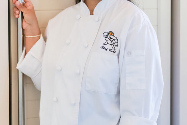 A woman from the Lotus Chefs team, Chef Belia Yolanda Paul, is smiling in her white chefs coat while standing by a louvered door. Her long dark hair is pulled back, and shes holding the door with one hand. The coat proudly displays an embroidered logo on the chest.