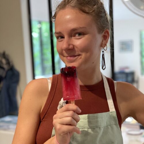 Chef Casia Rose from Lotus Chefs in a brown tank top and light green apron, a proud member of the Lotus Chefs team, holds a pink and purple popsicle with berries. She is indoors, smiling slightly, with a modern room and large glass windows in the background.