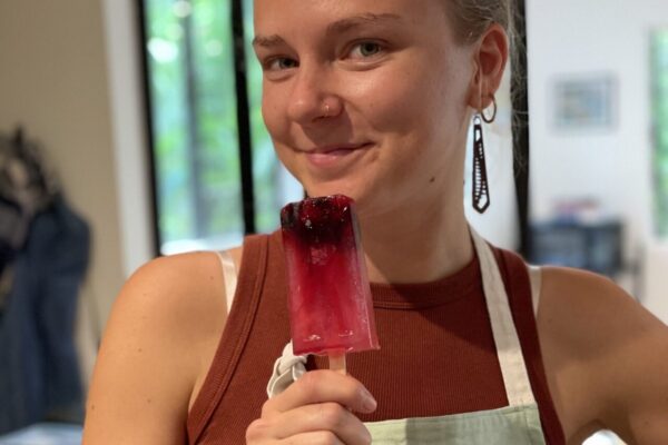 Chef Casia Rose from Lotus Chefs in a brown tank top and light green apron, a proud member of the Lotus Chefs team, holds a pink and purple popsicle with berries. She is indoors, smiling slightly, with a modern room and large glass windows in the background.