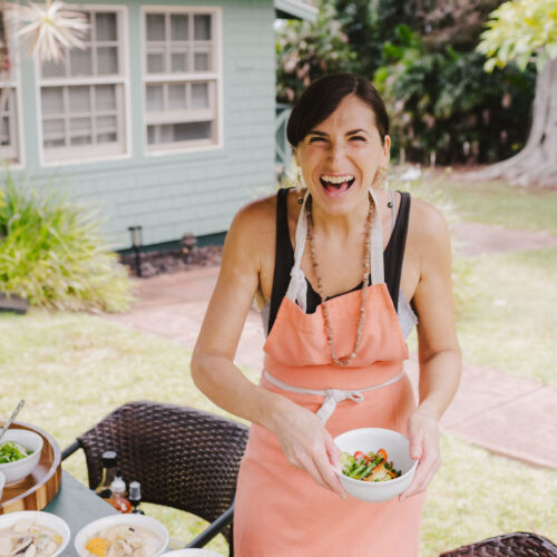 Chef Christina Oliveira from Lotus Chefs in an orange apron, part of the Lotus Chefs team, smiles broadly while holding a bowl of food outdoors. Various dishes are placed on a table beside her, with a green house and lush plants visible in the background.