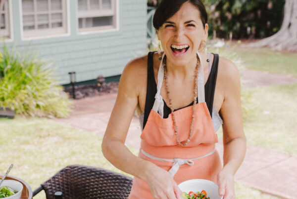 Chef Christina Oliveira from Lotus Chefs in an orange apron, part of the Lotus Chefs team, smiles broadly while holding a bowl of food outdoors. Various dishes are placed on a table beside her, with a green house and lush plants visible in the background.