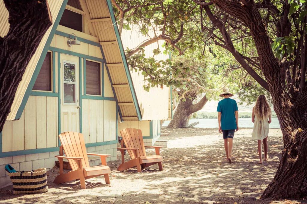 A man and a woman stroll on a sandy path beneath trees near an A-frame cabin, reminiscent of retreats at Camp Olowalu. Two wooden chairs sit outside, inviting relaxation. Nearby, a woven basket rests on the sand. The scene is sunny and tranquil.