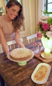 Chef Angel Green from Lotus Chefs smiles as she places a cake on a dining table, feeling like part of the lotus chefs team. The table is decorated with pink and white flowers, a lit candle, and glasses with a pink drink. Another dish with sliced fruit is also on the table.