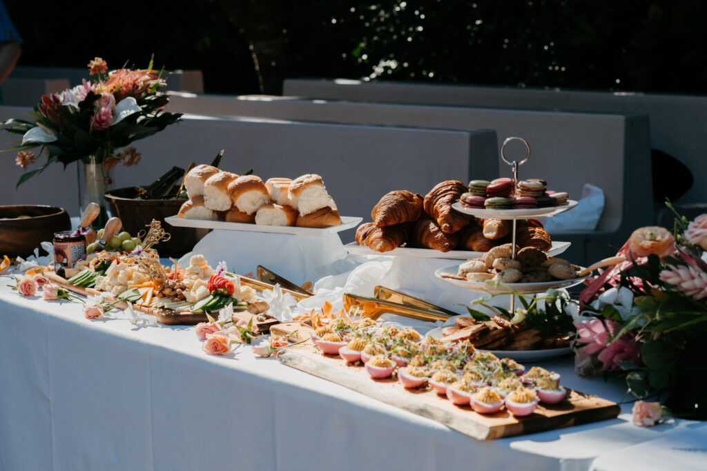 A buffet table is laden with assorted pastries, bread, macarons, and savory appetizers, featuring fresh florals as decoration for a brunch wedding private chef booking on Maui. Plates and serving utensils are neatly arranged on a white tablecloth in a bright outdoor setting.