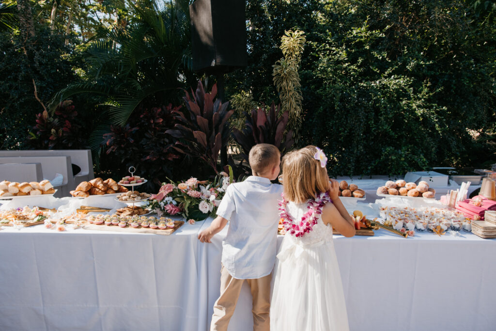 Two children stand at a styled grazing board buffet for a Maui wedding outdoors. The table is arrayed with pastries and finger foods. Both children are in formal attire, with the girl adorned in a floral lei. Tall greenery forms an enchanting backdrop, creating a picturesque setting for this elegant gathering.